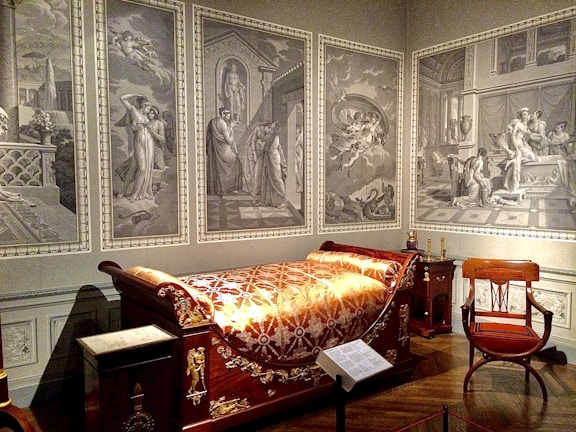 A room furnished with an opulent wooden bed adorned with gold patterns and an intricate blanket. The walls are decorated with large, monochromatic murals depicting classical scenes with figures in ancient attire. An elegant wooden chair with a red cushion is positioned near the bed, and various classical elements and decorations fill the room.