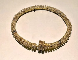 A circular gold necklace adorned with intricate, small, closely set embellishments creating a textured and ornate appearance. The piece reflects light, adding a lustrous quality to its design. It appears to be displayed flat against a neutral background, emphasizing its luxurious and opulent nature.