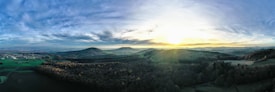 A stunning panoramic landscape showcases a serene countryside at sunrise. Rolling hills and dense forests stretch towards the horizon, with a glowing sun casting a golden hue over the scenery. Patches of green fields and a small village are visible in the distance. The sky is a blend of soft blue and grey clouds, creating a tranquil and expansive atmosphere.