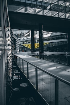 An urban architectural scene features a modern building complex with large glass windows and concrete structures. A prominent yellow truss adds contrast to the predominantly gray environment, while reflections on the water surface create intricate patterns. The geometric shapes, lines, and shadows contribute to a sense of depth and perspective.