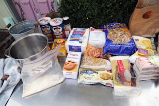 A collection of baking and cooking ingredients are displayed on a metal surface. These include a stainless steel pot, containers of oats, sugar, and flour, as well as packaged foods such as raisins, barley, and white chocolate. A paper bag and a metal bucket are also seen along with some soft fabrics.
