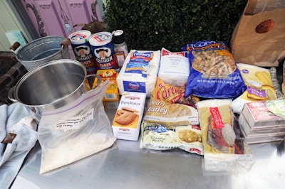 A collection of baking and cooking ingredients are displayed on a metal surface. These include a stainless steel pot, containers of oats, sugar, and flour, as well as packaged foods such as raisins, barley, and white chocolate. A paper bag and a metal bucket are also seen along with some soft fabrics.
