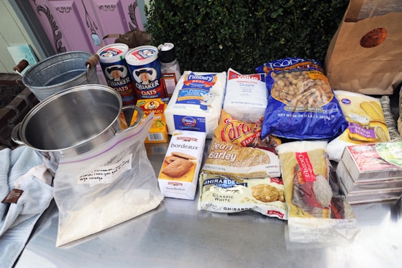 A collection of baking and cooking ingredients are displayed on a metal surface. These include a stainless steel pot, containers of oats, sugar, and flour, as well as packaged foods such as raisins, barley, and white chocolate. A paper bag and a metal bucket are also seen along with some soft fabrics.
