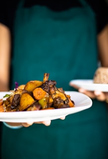 A person is holding two plates of food, one with a colorful assortment of roasted vegetables and meat, while the other plate appears to have a serving of grains. The person is wearing a teal apron, suggesting a culinary setting.