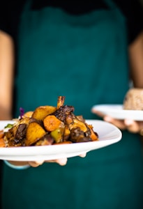 A person is holding two plates of food, one with a colorful assortment of roasted vegetables and meat, while the other plate appears to have a serving of grains. The person is wearing a teal apron, suggesting a culinary setting.