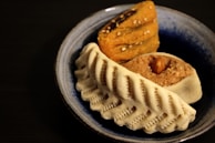 A blue ceramic dish contains an assortment of traditional sweets. The front item has a ridged white exterior, possibly a type of pastry or cookie. Behind it, a yellow-brown layered sweet sprinkled with seeds is visible. An almond is embedded on top of a beige-colored sweet.