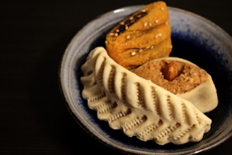A blue ceramic dish contains an assortment of traditional sweets. The front item has a ridged white exterior, possibly a type of pastry or cookie. Behind it, a yellow-brown layered sweet sprinkled with seeds is visible. An almond is embedded on top of a beige-colored sweet.