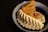 A blue ceramic dish contains an assortment of traditional sweets. The front item has a ridged white exterior, possibly a type of pastry or cookie. Behind it, a yellow-brown layered sweet sprinkled with seeds is visible. An almond is embedded on top of a beige-colored sweet.