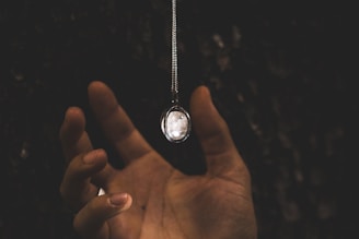 A hand is reaching out towards a pendant hanging from a chain. The background is dark, which makes the reflection on the pendant stand out. The overall lighting creates a moody atmosphere.
