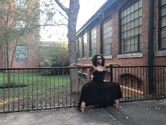 A person dressed in a black outfit balances gracefully in a dance pose on the railing of an outdoor path. The background features a brick building with large windows and a tree, surrounded by greenery.