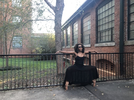 A person dressed in a black outfit balances gracefully in a dance pose on the railing of an outdoor path. The background features a brick building with large windows and a tree, surrounded by greenery.