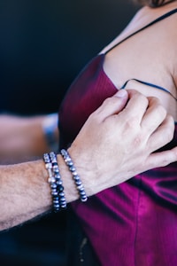 A person wearing a dark red dress is being assisted by another person who is adjusting the strap of the dress. The assisting person is wearing bracelets with black and white beads.