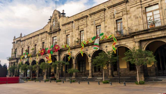 A historic stone building features a series of arched doorways adorned with colorful Mexican decorations, including green, white, and red garlands and festive ornaments. The building is characterized by detailed architectural elements and has a row of neatly trimmed trees in front of it.