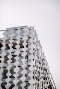 A modern building facade is adorned with a pattern of white geometric panels creating a visually striking and repetitive design. The panels are arranged vertically, attached to the exterior structure, adding a three-dimensional texture against a pale sky backdrop.
