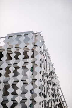 A modern building facade is adorned with a pattern of white geometric panels creating a visually striking and repetitive design. The panels are arranged vertically, attached to the exterior structure, adding a three-dimensional texture against a pale sky backdrop.