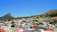 A vibrant neighborhood with brightly colored buildings nestled between two hills. The area boasts a variety of architectural styles and is rich in greenery, with trees scattered throughout. The sky is clear and blue, providing a picturesque backdrop to the lively scene.