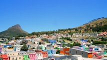 A vibrant neighborhood with brightly colored buildings nestled between two hills. The area boasts a variety of architectural styles and is rich in greenery, with trees scattered throughout. The sky is clear and blue, providing a picturesque backdrop to the lively scene.