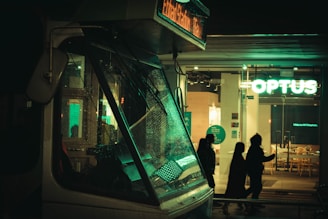 A street-level view at night features the front of a tram with a reflective windshield, illuminated by nearby lights. In the background, a brightly lit sign reads 'OPTUS' from a shop. Silhouettes of people are visible walking on the sidewalk, adding a sense of movement.