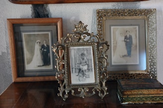 Three vintage photo frames are displayed on top of a wooden surface against a textured wall. The center frame, elegantly ornate, holds a photograph of a young child on a tricycle. The frames on either side contain sepia-toned wedding portraits of couples dressed in early 20th-century attire, featuring brides in long wedding dresses and grooms in formal wear. An antique-looking box is also present near the right frame.