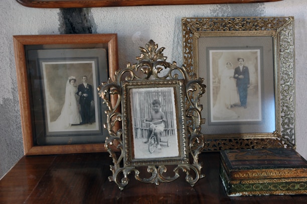 Three vintage photo frames are displayed on top of a wooden surface against a textured wall. The center frame, elegantly ornate, holds a photograph of a young child on a tricycle. The frames on either side contain sepia-toned wedding portraits of couples dressed in early 20th-century attire, featuring brides in long wedding dresses and grooms in formal wear. An antique-looking box is also present near the right frame.
