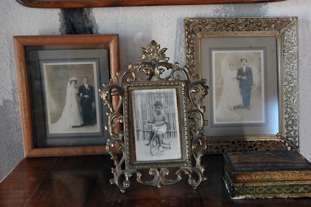 Three vintage photo frames are displayed on top of a wooden surface against a textured wall. The center frame, elegantly ornate, holds a photograph of a young child on a tricycle. The frames on either side contain sepia-toned wedding portraits of couples dressed in early 20th-century attire, featuring brides in long wedding dresses and grooms in formal wear. An antique-looking box is also present near the right frame.