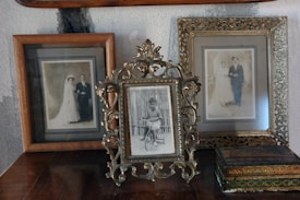Three vintage photo frames are displayed on top of a wooden surface against a textured wall. The center frame, elegantly ornate, holds a photograph of a young child on a tricycle. The frames on either side contain sepia-toned wedding portraits of couples dressed in early 20th-century attire, featuring brides in long wedding dresses and grooms in formal wear. An antique-looking box is also present near the right frame.