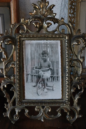 A black and white photograph of a young child sitting on a tricycle, framed in an ornate, decorative metal frame with intricate patterns. The background shows a wooden fence, suggesting an outdoor setting.