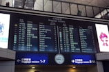 A digital airport baggage claim board displays flight information, showing flight numbers, departure cities, and corresponding baggage claim areas. The board is part of an airport terminal, with visible signage indicating directions to baggage claim areas. On either side of the board are advertisements, including a colorful cartoon character.