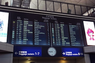 A digital airport baggage claim board displays flight information, showing flight numbers, departure cities, and corresponding baggage claim areas. The board is part of an airport terminal, with visible signage indicating directions to baggage claim areas. On either side of the board are advertisements, including a colorful cartoon character.