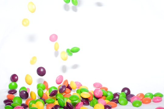 Brightly colored candies are scattered and appear to be falling against a white background. The candies are numerous and come in various colors including yellow, red, green, purple, and orange, creating a vibrant and lively scene.