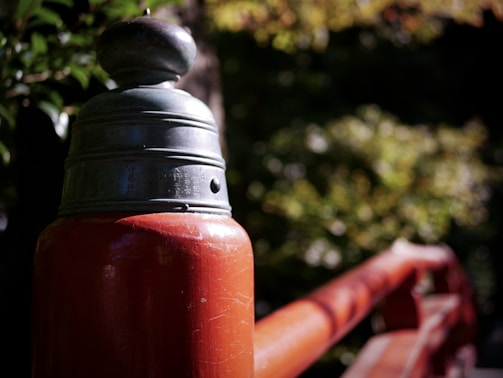 Close-up of a wooden railing with a decorative metal cap, featuring wooden elements painted in a dark red hue. The background displays out-of-focus leaves and branches, suggesting a natural setting with dappled sunlight filtering through.