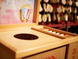 A wooden offering box typically found at Shinto shrines in Japan, featuring a round opening for coins. There are decorative plaques or charms hanging nearby, with a colorful board displaying small items. The background is slightly blurred, showcasing more traditional decor.