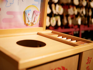 A wooden offering box typically found at Shinto shrines in Japan, featuring a round opening for coins. There are decorative plaques or charms hanging nearby, with a colorful board displaying small items. The background is slightly blurred, showcasing more traditional decor.