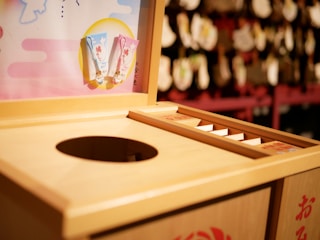 A wooden offering box typically found at Shinto shrines in Japan, featuring a round opening for coins. There are decorative plaques or charms hanging nearby, with a colorful board displaying small items. The background is slightly blurred, showcasing more traditional decor.