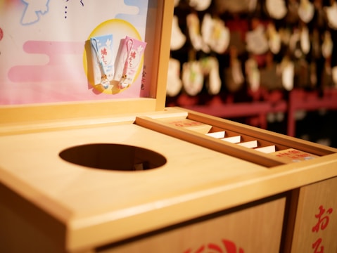 A wooden offering box typically found at Shinto shrines in Japan, featuring a round opening for coins. There are decorative plaques or charms hanging nearby, with a colorful board displaying small items. The background is slightly blurred, showcasing more traditional decor.