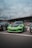 A vibrant green sports car is parked in front of a building with the sign that reads 'Ace Cafe Luzern'. The car is flanked by other vehicles and there are people gathered around the area, some of whom are seated at tables. The sky is overcast, adding a dramatic backdrop to the scene.