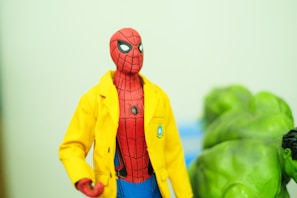 A figurine of Spider-Man wearing a bright yellow jacket is in focus. The background shows the partially visible figure of the Hulk, characterized by green skin and muscular build. The setting appears to be indoors with a soft greenish hue.
