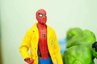 A figurine of Spider-Man wearing a bright yellow jacket is in focus. The background shows the partially visible figure of the Hulk, characterized by green skin and muscular build. The setting appears to be indoors with a soft greenish hue.