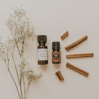 Two small bottles of essential oils are placed on a light surface, accompanied by several cinnamon sticks arranged to the right. A sprig of delicate white flowers lies on the left side, adding a natural touch to the composition.