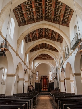 A spacious church interior with high arched ceilings and rows of wooden pews. The walls are adorned with tall, narrow stained glass windows, and chandeliers hang from the ceiling. The altar is situated at the far end of the aisle, framed by dark wooden paneling.