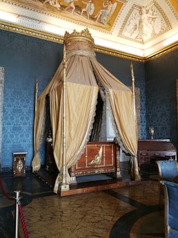 An ornate, vintage royal bed with a golden canopy and decorative drapes is placed in a lavishly decorated room. The room has dark blue walls, adorned with intricate patterns and a richly decorated ceiling featuring classical art. There is a wooden desk to the side and a chair with a blue upholstered seat. A red rope barrier is visible in the foreground.