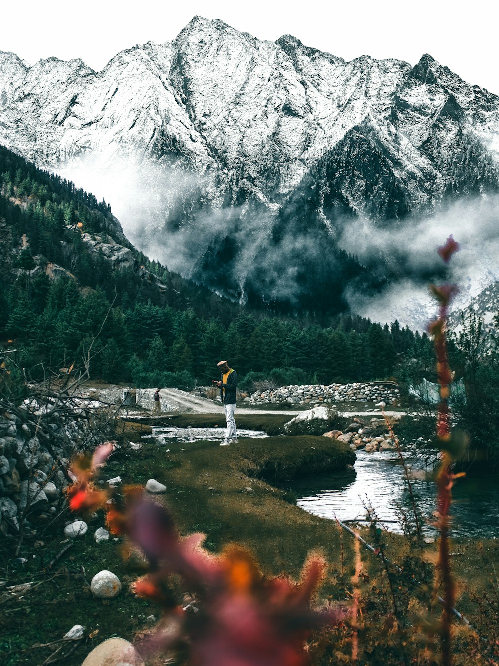 man standing near river overlooking snow capped mountains