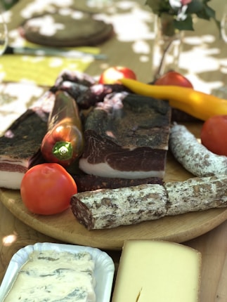 A wooden platter is adorned with an assortment of cured meats, including slices of salami and prosciutto. Fresh produce such as tomatoes and peppers adds vibrant color. There are also blocks of cheese, all set on a table with dappled sunlight filtering through leaves above.