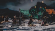 A detailed miniature wargame scene with well-crafted figures in armor holding weapons, set on a snowy terrain. The diorama includes small structures and gothic-style gates, suggesting a medieval or fantasy setting. The background shows green and dark tones that enhance the mysterious atmosphere.