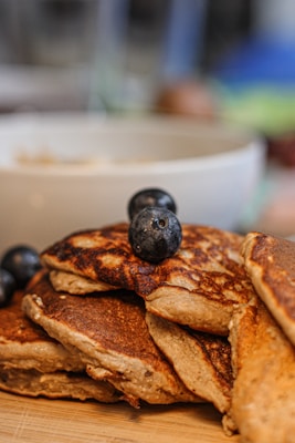 A close-up of a stack of fluffy pancakes topped with fresh blueberries. The pancakes have a golden-brown appearance and are placed on a wooden surface. A blurry background with a white bowl is visible, indicating a cozy breakfast setting.