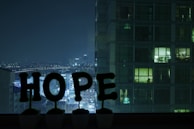 A nighttime cityscape view from a window, with large illuminated letters spelling out 'HOPE' placed on the sill. In the background, a high-rise building is partially visible with some lit windows. The city skyline is seen in the distance under a dark sky.