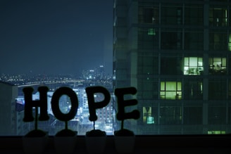 A nighttime cityscape view from a window, with large illuminated letters spelling out 'HOPE' placed on the sill. In the background, a high-rise building is partially visible with some lit windows. The city skyline is seen in the distance under a dark sky.