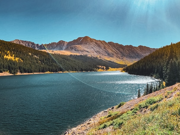 A serene mountain landscape features a clear blue lake surrounded by dense forests and majestic mountains in the background. The sunlight creates a shimmering effect on the water's surface.