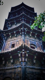 A traditional multi-tiered pagoda featuring intricate carvings and detailed architectural elements. The structure is made of stone, with each tier displaying ornate designs reminiscent of historical religious temples. Lush green tree leaves partially frame the top right corner, adding a natural contrast to the stone structure.