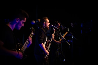 A group of musicians performing on stage, featuring a guitarist playing energetically in the foreground and a vocalist singing passionately into a microphone. The lighting is low, casting a dramatic and moody ambiance over the scene.